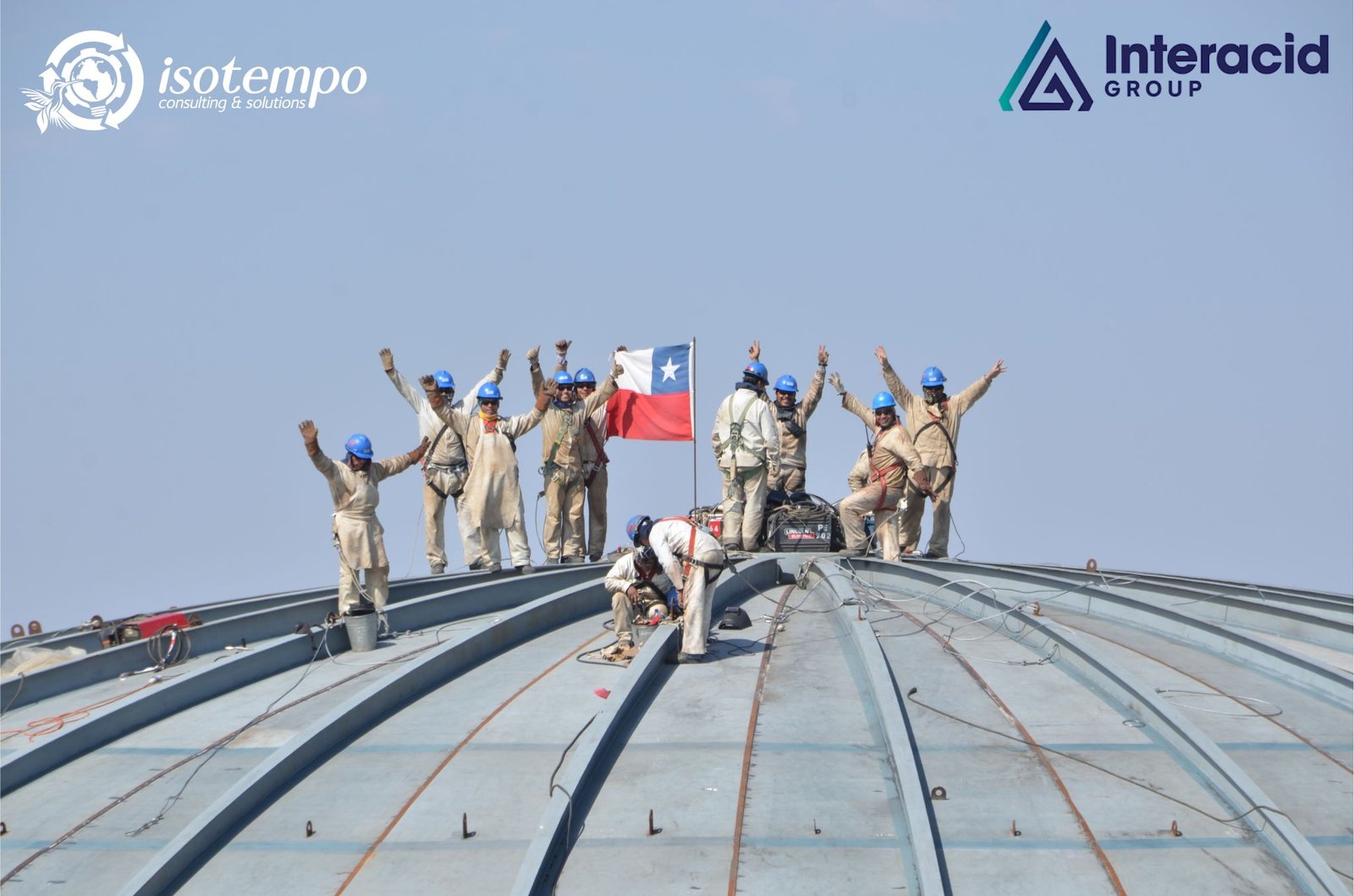 Trabajadores industriales con cascos azules celebrando sobre estructura metálica junto a la bandera de Chile – Isotempo Consulting & Solutions e Interacid Group